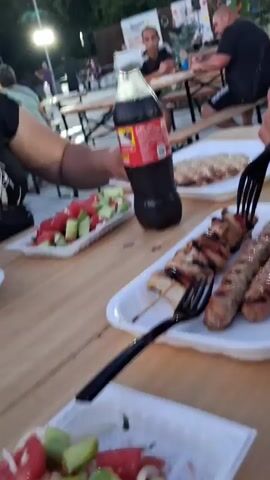 The video shows a group of people gathered at picnic tables outdoors. In the foreground, a woman in a black t-shirt is holding a fork with food and appears to be eating. A bottle of Coca-Cola is on the table next to her, along with a plate of salad containing tomatoes and cucumbers.

In the background, other people are seated at similar tables. A man in a black t-shirt is visible sitting and looking towards the left. Further back, a band appears to be setting up or playing on a stage with musical instruments. The overall scene suggests an outdoor event or gathering, possibly a festival or outdoor dining experience.

The time of day appears to be dusk or early evening, as indicated by the soft, ambient light and the clear, pale sky. The weather seems pleasant, as people are dining outdoors without apparent discomfort. The architecture of a building in the background, with its brick facade and windows, suggests an urban or semi-urban setting.

A sign with the word "Smart" and possibly other text in a foreign language is visible in the background, though it is difficult to read clearly. The general atmosphere seems relaxed and social, with people enjoying food and company.