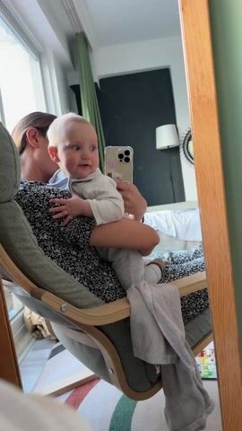 A woman sits in a rocking chair with a baby in her arms, looking into a mirror. The woman wears a dark floral dress and has long blonde hair pulled back. The baby wears a light grey onesie and looks at the camera. The woman holds a cell phone, presumably taking a selfie. The rocking chair is light green with a wooden frame. The room is bright with natural light coming through a window to the left of the frame. Behind the woman and baby, a crib is visible against a dark grey wall. There is a green curtain near the window and a colorful rug on the floor. A lamp and a decorative wall mirror are also visible in the reflection. The woman kisses the baby on the cheek.