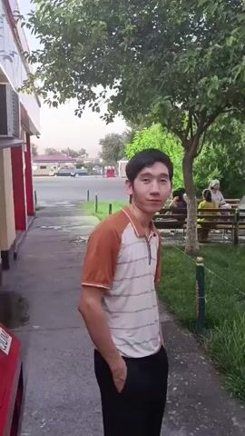 The video shows a young man standing next to a sidewalk. He is wearing a striped polo shirt and dark pants. He is looking at the camera and smiling. There are trees and benches in the background, and other people are sitting on the benches. The scene appears to be outdoors, possibly in a park or a public area.