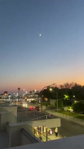 The video shows a twilight scene over a parking lot and a street with a few cars and streetlights. In the distance, there is a body of water with some buildings and palm trees along the waterfront. The sky is a gradient of blue and orange, with a crescent moon and a bright star visible. There are several people walking in the foreground near a building entrance. The camera pans from left to right, capturing the expanse of the parking lot filled with vehicles and the silhouetted trees against the colorful sky.