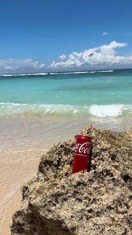 The video shows a red Coca-Cola can perched on a textured rock near a sandy beach. The ocean is in the background, displaying a turquoise color with white waves rolling in. The sky is blue with scattered white clouds. The overall scene depicts a sunny day at the beach. No people are visible.