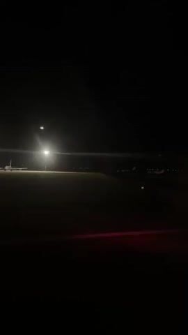 This is a low-light video from inside an airplane cabin, likely taken from a window seat. The plane is on the tarmac at night, with runway lights visible in the distance. A bright spotlight illuminates a portion of the runway. A few other planes or ground vehicles are faintly visible further down the tarmac. The red lights of the runway are also visible. The plane appears to be moving slowly.