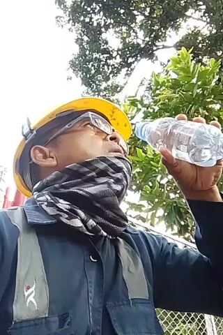 A man wearing a yellow hard hat, safety glasses, and a patterned neck gaiter is drinking from a clear plastic bottle of water. He is wearing a dark blue shirt or jacket with a gray vest over it. He is holding the bottle in his right hand and tilting his head back to drink. In the background, there are green trees and a wire fence. The sky is visible above the trees.