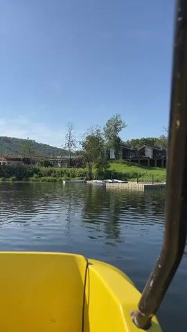 The video shows a view from the front of a bright yellow paddle boat on a calm lake under a clear blue sky. The boat is moving forward, indicated by the water rippling around the bow. The shoreline is visible in the background, with lush green trees and rolling hills. There are buildings on the far shore, some with dark exteriors and large windows, nestled amongst the trees. A small dock or platform juts out into the water. Further out on the lake, other paddle boats can be seen, and at least one person is visible walking on the shore near the buildings. The sun is shining brightly, casting reflections on the water and creating a bright, sunny atmosphere.