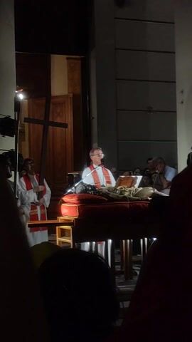 The scene depicts a religious ceremony taking place in what appears to be a church or cathedral. A man wearing a white shirt with a red and white stole is standing at a podium, addressing an audience. He is illuminated by a spotlight, highlighting him and the podium where a figure resembling a deceased Christ is laid on a red cushion. 

To the left of the speaker, two other individuals dressed in white robes and red stoles are standing. One of them is holding a large wooden cross. The lighting is dim, creating a solemn atmosphere. The audience is visible in the background, and details of the church's interior are visible, including a large wooden door behind the speakers and stone walls. The video seems to be shot from within the audience, partially obscured by the heads of people in front.
