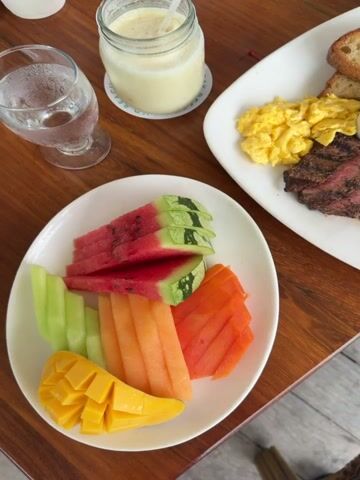 A video shows a close-up overhead view of a wooden table with a meal laid out. In the foreground, a white plate contains sliced watermelon, cantaloupe, and mango. To the right, a larger white plate holds scrambled eggs, sliced steak, two potato pancakes, two pieces of toast, a small ramekin of sauce, and a small ramekin of butter. To the left, a glass of water with ice is visible, and behind that, a glass jar with a yellow beverage and a straw. The lighting is natural, suggesting an outdoor or well-lit indoor setting. There are no people visible in the video.