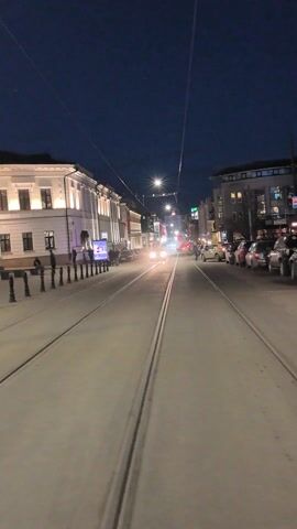 The video shows a street scene at night, likely in a city. The camera moves from an empty street with tram tracks to a pedestrianized area filled with people. The buildings on either side of the street are lit up. There are streetlights and signs. People are walking in various directions, some in groups. The overall environment appears to be a public space for leisure or commuting.