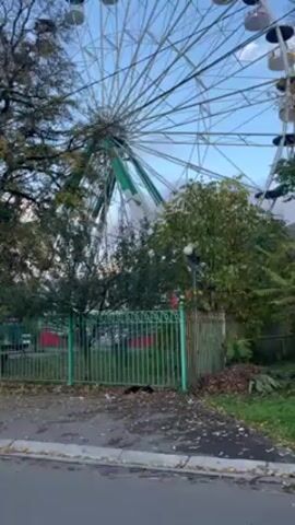 The video shows a Ferris wheel behind a green fence in what appears to be a park, then pans to show an empty street leading uphill, with trees lining the sides, under a blue sky with scattered clouds.
