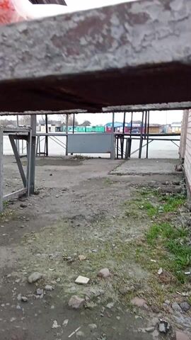 The camera moves through an empty outdoor market under a metal awning with open sides. There are metal framed tables but no vendors. The ground is cracked asphalt with loose gravel and dirt. The camera passes along the back of a white brick building with broken portions of brick walls.  A white plastic bag moves in the top right of the screen. The sky is overcast.