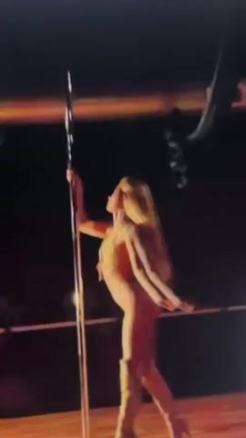 A blonde woman in a shiny gold outfit is performing a pole dance. She is holding onto a vertical pole with her hands and has her legs wrapped around it. She is rotating around the pole and her hair is flying around her head. She is wearing high heeled boots. The background is dark, with some lighting illuminating the stage. There are some draped fabric elements hanging from the ceiling in the background.