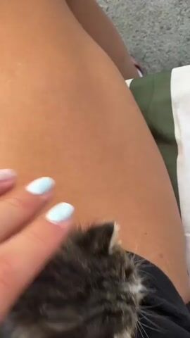 A close-up shot shows a person's hand with light blue painted fingernails and a ring on their finger gently stroking a very small, fluffy kitten. The kitten is mostly gray and white and appears to be lying on a person's lap, possibly on black fabric shorts. The background includes the person's tanned thigh and a striped fabric, possibly a blanket or cushion, with green and white stripes. The kitten is the primary focus, and the interaction between the hand and the kitten suggests a moment of comfort or play.