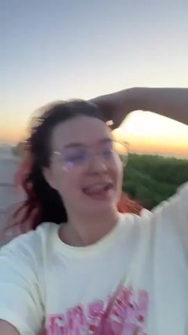The video shows a young woman with dark hair, some of which appears to be dyed a reddish-pink, wearing glasses and a white t-shirt with pink text. She is posing for a selfie against a sunset sky. The sky is a gradient of soft blues and oranges. In the background, there is a blurry field of green grass or reeds. The woman is smiling and making playful faces at the camera. The scene suggests a casual, outdoor setting during the golden hour. The visible text on her shirt reads "MONSTER" in large, stylized pink letters, with smaller text underneath that is difficult to decipher but appears to be playful or stylized. The overall mood is cheerful and relaxed.