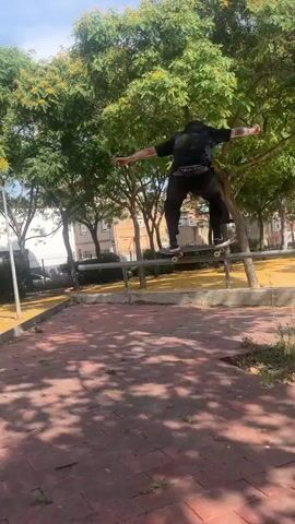 A male skateboarder is performing a trick on a metal rail in an outdoor park. He is wearing dark clothing and is in mid-air with his skateboard. In the background, another male is standing and holding a phone, possibly filming. Trees with green leaves and scattered yellow leaves are visible, along with buildings and a fence. The ground is a mix of reddish-brown pavers and sandy dirt. The overall scene depicts a sunny day in a park setting where skateboarding is taking place.