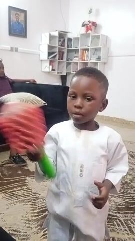 A young Black boy wearing a long white shirt and pants is standing in a living room while holding a handheld rattle. He is looking directly at the camera while shaking the rattle. A man is sitting on a black couch in the background. There is a geometric shelf unit behind the couch. The room has a patterned rug. There is a framed photo on the wall.