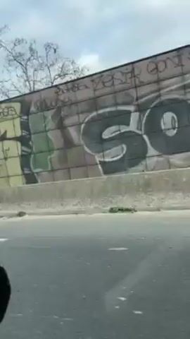 The video shows a view from inside a car traveling on a highway. The camera is positioned behind the dashboard, facing forward.  The outside environment, seen through the car window, includes a grey cloudy sky, and roadside elements. On the right of the road, there's a barrier with graffiti art. As the car moves, the view extends to show a bridge structure with bare trees and buildings in the background. The road surface is asphalt with white lane markings. The dashboard contains items such as a small pouch. The video continues with the car moving down the highway.