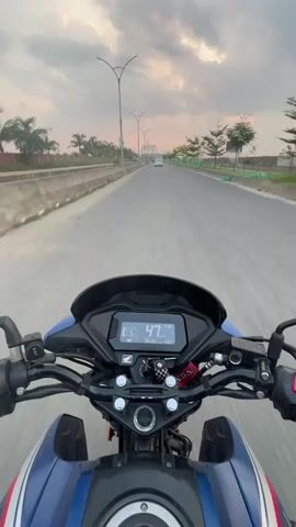 The video captures a first-person perspective of someone riding a blue Honda motorcycle on a multi-lane road in Rupganj, Bangladesh. The camera is mounted on the motorcycle, providing a clear view of the handlebars, dashboard, and the road ahead.

The dashboard, visible in the foreground, shows a digital speed reading that fluctuates from 48 km/h to 54 km/h. The gear indicator displays '5', and the odometer reads 10393.3 km. A "HONDA" logo is prominently displayed on the motorcycle's tank, and a red and white keychain hangs near the ignition.

The road ahead is straight and appears well-maintained, flanked by concrete barriers, palm trees, and other young trees. Streetlights with multiple arms line both sides of the road, extending into the distance where a bridge or overpass is visible. Other vehicles, primarily cars, can be seen in the far distance.

The sky is largely covered with clouds, but soft, diffused sunlight, possibly from late afternoon or early evening, creates a gentle glow, particularly on the horizon. The overall impression is of a calm ride on a relatively open road during dusk or dawn.