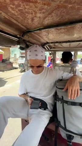 A person wearing white clothes, sunglasses, and a white bandana sits in the back of an auto-rickshaw, holding a phone. A grey backpack sits next to the person. The auto-rickshaw moves along a road in India. Motorbikes, trucks, and other vehicles pass by. The rickshaw has a weathered, rusty roof.