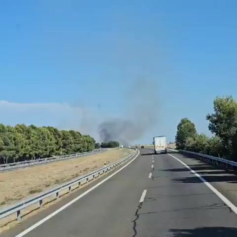 A first-person perspective video shows a view from a moving vehicle on a highway. The vehicle is traveling on the right side of the road. The sky is clear and blue. On the left side of the highway, there is a guardrail and then dry, grassy terrain with scattered trees. On the right side of the highway, there is a similar guardrail and terrain, with more trees visible in the distance. Ahead, a large plume of dark smoke is rising into the sky, obscuring the horizon. Several other vehicles, including a white truck, a white van, and a few cars, are visible on the highway, also moving away from the smoke. The smoke appears to be coming from a fire in the distance, off to the left of the highway.