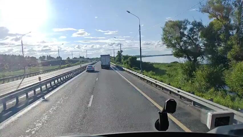 The video shows a first-person view from inside a vehicle driving on a highway. The highway has multiple lanes separated by yellow lines. On the left side of the highway, there is a metal guardrail, and beyond that, more lanes of traffic with a truck and a white car visible. On the right side of the highway, there is also a guardrail, and beyond that, lush green grass and trees line the edge of a large body of water, possibly a river or lake. The sky is blue with scattered white clouds, and the sun is shining brightly, casting a glare on the windshield. A black object, possibly a dashcam mount, is visible in the lower part of the windshield. Several lampposts are visible along the side of the highway. The overall scene suggests a clear, sunny day on a highway in a rural or semi-rural setting with water nearby.