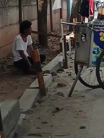 A young man in a white t-shirt and dark pants sits on the ground next to a concrete wall and a sidewalk. He is looking to his left. Next to him, on the sidewalk, is a food cart with two wheels. The cart has a metal exterior and appears to be selling some kind of food or drink, with bottles and containers visible on top. The cart has signage with text and images on it, including the words "Sari Rasa" prominently displayed. The ground is a mixture of dirt and gravel. Trees are visible behind the man and the cart. In the background, there are other carts, some parked motorcycles, and people walking. The scene appears to be outdoors, possibly on a street or market.