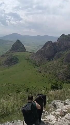 The video shows two people wearing backpacks and descending a rocky hill towards a green valley. A third person wearing an orange hat joins them and they all look out over a landscape of rolling green hills, a uniquely shaped triangular peak, and distant mountains beneath a sky with puffy clouds.