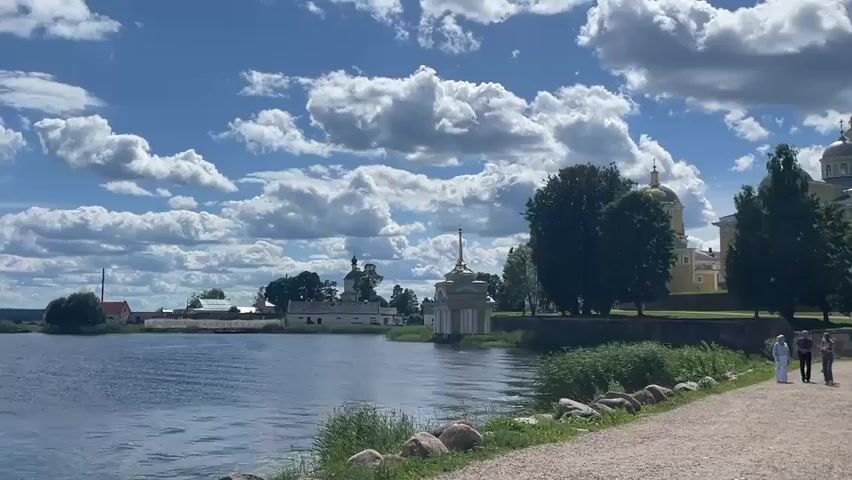 The video shows a scenic view of a body of water with a pathway leading towards a complex of buildings, possibly a monastery or cathedral. The sky is partly cloudy with dramatic white clouds against a bright blue background. On the right side of the frame, there is a pathway lined with rocks and green vegetation. A few people are walking on the pathway, appearing to be tourists. In the background, there are several buildings with yellow walls and golden domes, indicative of religious architecture. Trees are scattered throughout the landscape. On the left side of the water, there are more buildings, some with green domes and towers, and a distant fence. The water appears calm and reflects the sky and surrounding environment. The overall atmosphere is peaceful and picturesque, suggesting a summer day.