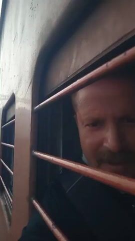 The video shows a man with fair skin and a mustache looking out a train window. He is wearing a black jacket with a white fur collar. The train window has orange bars. The man smiles and laughs. Outside the window, train tracks and a misty landscape are visible. The train appears to be moving. Another person is faintly visible behind the man.
