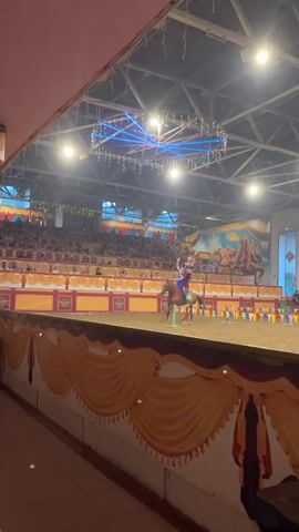 A horse show is taking place indoors.  The horse is brown and is being ridden by a person in a blue and red costume. Another person is standing on the rider's shoulders, also in a costume. The horse is moving across a dirt arena. There are spectators seated in tiered seating in the background. Colorful banners and decorations are visible. The ceiling has lights and a decorative structure with lights. Another horse and rider are in the background.