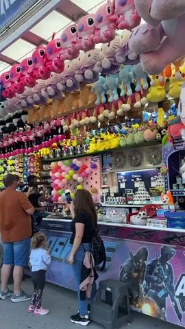 The video shows an outdoor carnival game booth filled with numerous stuffed animals hanging from the ceiling. The booth has a colorful backdrop with a balloon game in the center. Several people are gathered around the booth, including a man, a woman, and a young child, who appear to be playing or watching the game. Another person is behind the counter, likely operating the game. The atmosphere seems lively and festive, characteristic of a fair or carnival.