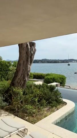 The video shows a tranquil scene of an outdoor swimming pool overlooking a bay or sea. The camera is positioned under a roof or overhang, providing a partially framed view. In the foreground, there's a large tree trunk and surrounding greenery, creating a natural barrier between the camera and the pool area. Several lounge chairs are visible next to the tree. The pool itself has a modern, geometric design with light-colored stone trim. The water is clear and reflects the surrounding environment. Beyond the pool, there's a narrow strip of beach, green bushes, and several white umbrellas providing shade. The body of water in the background is dotted with boats, including a sailboat, and a coastline is visible in the distance under a slightly cloudy sky.