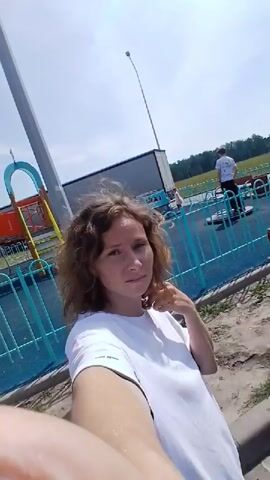 A woman with curly brown hair and a white t-shirt stands in front of a playground. She is holding the camera, taking a selfie. Behind her, there is a blue fence, a slide, and a truck. The sky is blue and sunny. She turns her head to the right, then to the left. The camera pans slightly to the right, showing a building in the background with people walking in front of it.