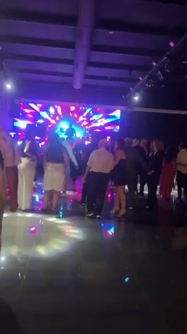 A group of people are dancing in a dimly lit room with a large screen displaying abstract, colorful patterns in the background. The lighting is predominantly purple, blue, and green, with some yellow and pink accents. The floor is dark and reflective, showing the lights and the people's reflections. In the foreground, several individuals are visible, some dancing together, others standing and watching. One woman in a sparkling gold outfit appears to be singing or speaking into a microphone. The atmosphere seems festive, possibly a party or a wedding reception.