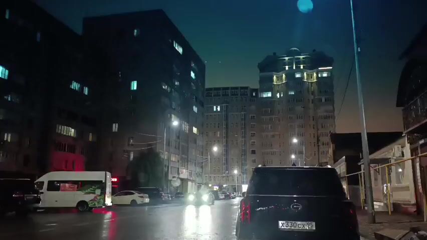 The video shows a nighttime view from inside a vehicle driving on a wet city street. Tall buildings with illuminated windows line both sides of the street. Headlights from oncoming and parked cars are visible, reflecting on the wet asphalt. The video captures the rear of the vehicle the camera is in, a black SUV with a license plate visible. Traffic lights and streetlights are also present.