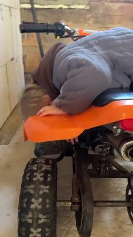 A person is lying on top of a quad bike. The person is wearing a gray jacket over a brown hoodie and has blonde hair. The person's face is buried in the seat of the quad bike, and their hands are near the rear wheel. The quad bike is orange and black, with large tires. The background appears to be a garage or workshop, with a concrete floor and a brick wall. The lighting is dim. The video is very short, with rapid cuts between slightly different angles.