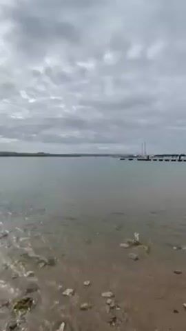 The scene shows a body of water, possibly a bay or inlet, under a cloudy sky. The water is calm with gentle ripples, and the shoreline is visible in the foreground with rocks and sand. In the distance, a landmass is visible along the horizon. There are boats or masts discernible in the water further out. The camera is static. The lighting is subdued due to the overcast sky. The sun peeks out for a moment.