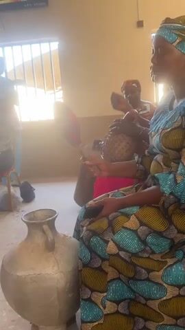 A low-angle shot focuses on two women sitting on a bench in a room filled with sunlight. The woman in the foreground is wearing a colorful, patterned dress and is positioned to the right of a large, stone-like vessel. She holds a long stick with a red, pom-pom-like end, rhythmically striking the top of the vessel. The second woman is seen in the background wearing an orange patterned shirt and sits with an ornate basket on her lap. A window with bars is visible in the background, with light streaming in. The camera remains static, focusing on the action with no cuts or zooms.