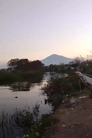 A wide shot captures a serene natural landscape at dusk. A still body of water occupies the left half of the frame, its surface reflecting the soft hues of the twilight sky. Lush green foliage lines the banks of the water, with trees exhibiting autumnal shades of red and brown. In the distance, a majestic mountain rises, its peak softly illuminated by the fading sunlight. The right side of the scene depicts a more rugged, unkempt area with scattered debris and vegetation. A rough, dirt path runs along the edge of the water, suggesting a rural or less developed setting. The overall atmosphere is peaceful and tranquil, with a sense of natural beauty dominating the visual narrative.