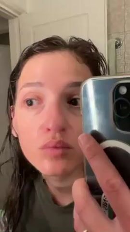 A woman with wet dark hair stands in a bathroom holding a silver iPhone in her right hand, pointed at herself. Her face is close to the camera, showing her eyes, nose, and mouth. She sticks her tongue out and moves her eyes, looking towards the left. A white door is visible in the background, leading to what appears to be a shower or tub area with patterned tile. The bathroom mirror reflects her image.