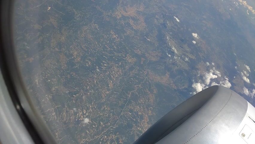 This media file shows an aerial view from an airplane window, looking down at the earth. The primary subject is the landscape below, which appears to be a mix of rural and natural terrain. We can see winding lines that suggest rivers or roads, and patches of different colors indicating fields or vegetation. There are also scattered clouds in the sky. A portion of the airplane's engine nacelle is visible in the upper left corner of the frame, indicating the perspective is from inside the aircraft.

The scene is consistent with daytime, and the weather appears to be partly cloudy. The view suggests the plane is flying over a landmass, likely a continent or large island, given the detailed view of the terrain. There are no people visible in the media file. The overall mood is one of travel and observation of the world from a distance. No text is discernible within the frame. The provided location context of Ilidža, Bosnia and Herzegovina, suggests this is the geographical region being viewed, although specific landmarks are not identifiable from this altitude and perspective.