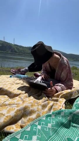 The video shows two people enjoying a sunny day outdoors by a large body of water, likely a river, with green hills in the background.

In the first shot (0-1 second), one person is seen sitting on a yellow patterned blanket spread on the grass. They are wearing a black bucket hat, a pink and white plaid shirt, and dark pants, and are actively using a silver laptop. A plastic water bottle lies beside them. Power lines are visible on the distant hills under a clear blue sky.

The second shot (1-5 seconds) changes perspective, focusing on the propped-up legs and feet of another person. This person wears light grey cargo pants and distinctive colorful sneakers (mint green, pink, and black). In the background, the first person, or a companion, is seen bending over near the water's edge, appearing to prepare a grill or fire pit. A camping chair and some bags are on the ground near them. Lush green grass and trees frame the scene, emphasizing the natural outdoor setting. The weather remains bright and clear throughout the video, suggesting a pleasant day in Sliznevo, Russia.