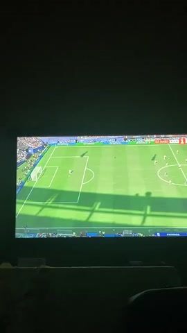 The video shows a television screen displaying a soccer game. The game is being played on a green field with white lines. There are players on the field, and a soccer ball. The game appears to be in progress. The screen is large and the image is clear. The lighting in the room is dim, with shadows cast on the screen from an unseen object.