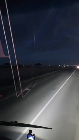The media file shows a first-person perspective from inside a vehicle driving on a road at dusk or night. The headlights of the vehicle illuminate the asphalt road ahead, which has a white line dividing the lanes. In the distance, other vehicles with their headlights on can be seen. The sky is dark blue with some clouds, and there are lights from a distant town or settlement visible on the horizon. The scene appears to be rural or on the outskirts of a town. A dash camera is visible in the foreground, mounted on the windshield. There are also some wires or cables running across the windshield, possibly from the vehicle's interior. On the right side of the road, a speed limit sign is visible, indicating "30". The overall mood is calm and perhaps a bit somber due to the time of day and the vast, open road. The text "АКБ" is visible on a piece of paper or label in the foreground.
