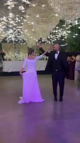 A wedding dance scene features a bride and groom under elaborate chandelier-like lighting. The bride wears a white, floor-length dress with three-quarter sleeves and has her hair up. The groom is in a black tuxedo with a bow tie and a neatly trimmed beard. They perform a dance move with their arms outstretched, palms facing up. Behind them, guests are seen clapping and watching. The room is decorated with a long white table, greenery, and mirrored accents. The dance floor is a light, polished surface. The atmosphere is celebratory.
