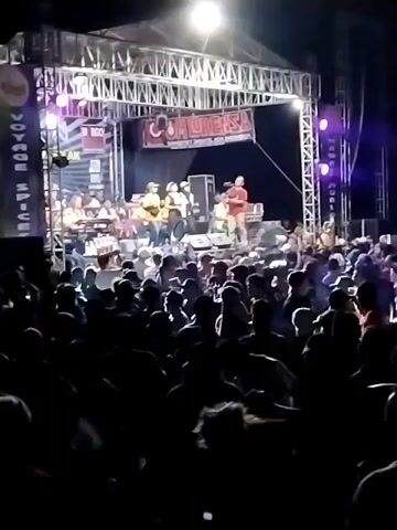 The video shows a nighttime outdoor concert with a live band performing on a stage. A crowd of people is gathered in front of the stage, some of whom are silhouetted against the stage lights. The stage is set up with lighting rigs and banners displaying text, including what appears to be "COMMUNALISA". The band members are visible on stage, and some appear to be singing and playing instruments. The crowd is dense and appears to be actively engaged with the performance. The overall environment is dark, illuminated by the stage lighting and some ambient lights. The video quality is low, with a blurry and grainy appearance.