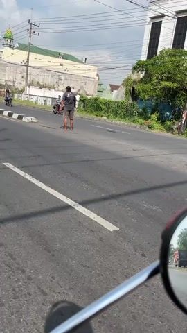 The video shows a street scene with a person standing in the middle of the road. The person is wearing a dark t-shirt and shorts. They appear to be interacting with a motorcycle. In the background, there are buildings, trees, and a mosque. The sky is visible with some clouds. The video is shot from the perspective of someone on a motorcycle, indicated by the handlebar and side mirror in the foreground. The road is paved and appears to be a city street.