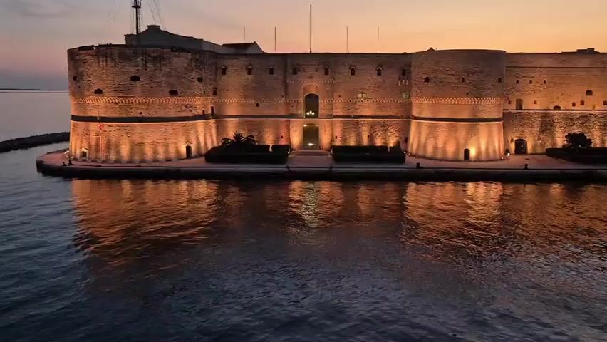 This is an aerial drone shot of Aragonese Castle in Taranto, Italy, at dusk. The ancient stone fortress, illuminated by warm spotlights, sits majestically on a promontory, surrounded by the calm, dark waters of the sea. The castle's imposing walls and battlements are clearly visible, with its reflection shimmering on the water's surface. The sky above is a gradient of soft orange and purple hues, typical of a sunset. 

The scene evokes a sense of historical grandeur and tranquility. The lighting creates a dramatic and inviting atmosphere, highlighting the architectural details of the castle. No people are visible in the frame, emphasizing the solitary and timeless nature of the structure. The weather appears calm, with no discernible wind or precipitation. The presence of a small breakwater made of large rocks to the left of the castle provides a subtle clue to its maritime setting. The overall mood is peaceful and awe-inspiring, inviting viewers to contemplate the history and beauty of this iconic landmark. There is no visible text in the media file.