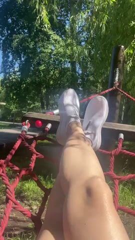 A first-person perspective video shows a person's legs and feet from the knees down. The person is wearing white slip-on sneakers and is sitting on a red rope net structure, possibly part of a playground. Their legs are crossed at the knees. The background consists of lush green trees and dappled sunlight, suggesting an outdoor, possibly park-like setting. There is a wooden beam and some red rope with metal fasteners visible in the foreground, to which the net is attached.