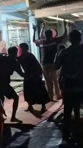 The video captures a group of people gathered in what appears to be an outdoor setting at night. The lighting is dim, with artificial lights illuminating the scene. In the foreground, a person in dark clothing is bent over, seemingly engaged in an action with another person. The second person is also in dark clothing and has their arms raised. Behind them, several other individuals are visible. One person in a striped shirt has their arms raised in the air. Another person is holding a pink balloon. The ground is covered with a patterned rug or mat, and there are other objects on the ground, including what looks like plastic sheeting. The background shows structures that resemble buildings with windows and a corrugated roof. The overall atmosphere suggests a social gathering or celebration.