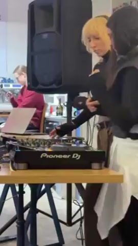 The video shows two people DJing at a cafe or restaurant. The person on the left has short blonde hair and is wearing headphones, a black top, and brown pants. They are using a Pioneer DJ mixer. The person on the right has long dark hair and is wearing a black vest over a white skirt. There is a large speaker next to them. A third person is sitting in the background using a laptop and a cell phone. The cafe has tables and chairs. The music is loud with a strong beat.