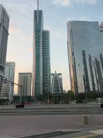 The video shows a cityscape with several modern skyscrapers. The buildings are tall and made of glass and steel, reflecting the sky and surrounding environment. There is a street in the foreground with a red pedestrian walkway and a roadway. People are walking along the pedestrian walkway, and cars are driving on the roadway. A bright yellow Lamborghini Urus drives past. The sky is blue with some clouds, and the overall lighting suggests it is daytime, possibly early morning or late afternoon. The camera seems to be panning from left to right, following the car. The video appears to have been taken from a moving vehicle.